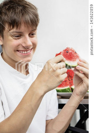 Cheerful young man winking and smiling while holding a fresh juicy watermelon piece. Tasty summer fruit snack on the table. Cheerful young man winking and smiling while holding a fresh juicy watermelon piece. Tasty summer fruit snack on the table. 141622859