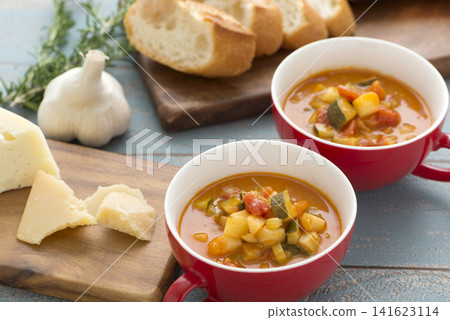 A table setting with warm minestrone in a red soup cup and baguette. A table setting with warm minestrone in a red soup cup and baguette. 141623114