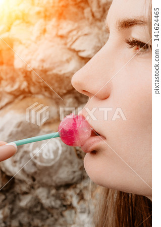 Woman lollipop close-up portrait of a young girl eating a pink candy outdoors against a natural rock background with sunlight Woman lollipop close-up portrait of a young girl eating a pink candy outdoors against a natural rock background with sunlight 141624465