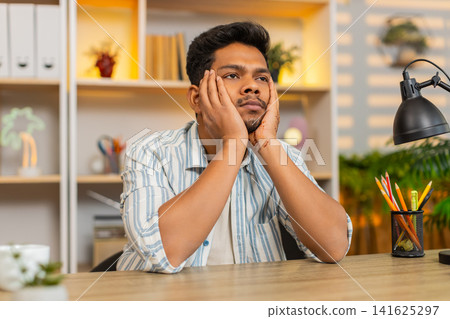 Indian man on home table leaning on hands looking exhausted after long remote work indoors at home Indian man on home table leaning on hands looking exhausted after long remote work indoors at home 141625297