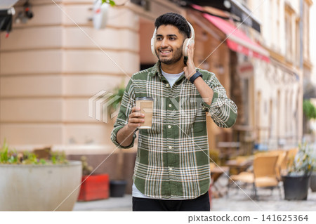 Indian man listening to music walking in city street with wireless headphones smiling during stroll Indian man listening to music walking in city street with wireless headphones smiling during stroll 141625364