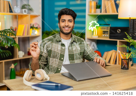 Indian man on home table closing laptop after work removing glasses with satisfied smile to camera Indian man on home table closing laptop after work removing glasses with satisfied smile to camera 141625418