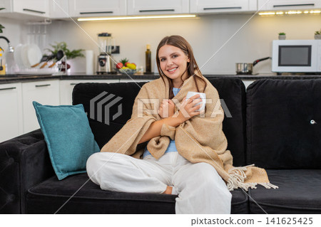 Young woman drinks herbal tea holds mug takes slow sip wraps in blanket relaxes and enjoys warm rest Young woman drinks herbal tea holds mug takes slow sip wraps in blanket relaxes and enjoys warm rest 141625425