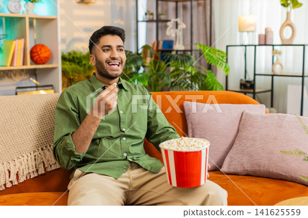 Indian man on home sofa watching movie with popcorn laughing and pointing during fun film night joy Indian man on home sofa watching movie with popcorn laughing and pointing during fun film night joy 141625559