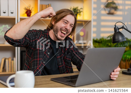 Young man on home table celebrating success with laptop after seeing great online result on screen 141625564