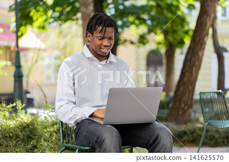 African American young man opening laptop for remote work in city street park outdoor urban travel African American young man opening laptop for remote work in city street park outdoor urban travel 141625570