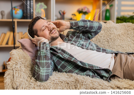 Young man on home sofa meditating with wireless headphones and calm smile during quiet music break Young man on home sofa meditating with wireless headphones and calm smile during quiet music break 141625608