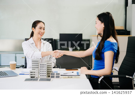 Asian female professionals shaking hands over a miniature building model during a real estate project or property investment agreement. Asian female professionals shaking hands over a miniature building model during a real estate project or property investment agreement. 141625937