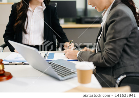 Two professional Asian female attorneys discussing legal documents and using a laptop in a modern law office workspace. 141625941