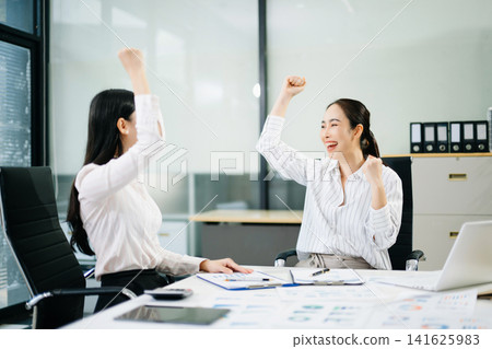 Two cheerful professional female colleagues raise their fists in celebration of a business achievement, major deal, or financial Two cheerful professional female colleagues raise their fists in celebration of a business achievement, major deal, or financial 141625983