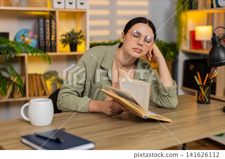 Young woman on home table reading book feeling sleepy during studying with glasses at desk indoors Young woman on home table reading book feeling sleepy during studying with glasses at desk indoors 141626112