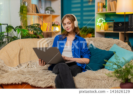 Young woman on home sofa working on laptop in headphones with focused modern freelancer mood indoors Young woman on home sofa working on laptop in headphones with focused modern freelancer mood indoors 141626129