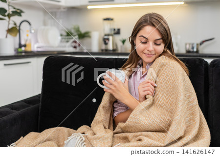 Young woman holds tea mug warms hands smiles under blanket enjoys quiet pause and peaceful deep rest Young woman holds tea mug warms hands smiles under blanket enjoys quiet pause and peaceful deep rest 141626147