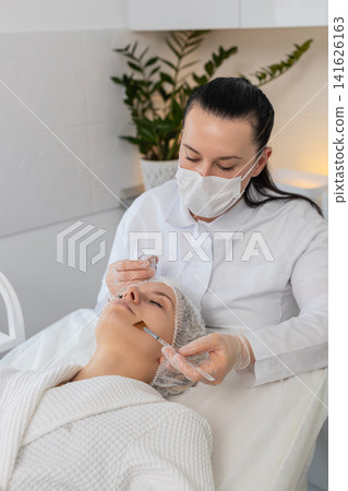 Enzyme therapy, beautician applying moisturizing medical cream on young woman face with brush Enzyme therapy, beautician applying moisturizing medical cream on young woman face with brush 141626163