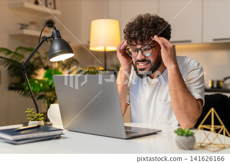 Arabian man holds head from screen strain takes break then resumes laptop work evening indoors Arabian man holds head from screen strain takes break then resumes laptop work evening indoors 141626166