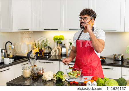 Arabian young adult man mixing salad and talking on phone in kitchen sharing healthy diet fit recipe Arabian young adult man mixing salad and talking on phone in kitchen sharing healthy diet fit recipe 141626167