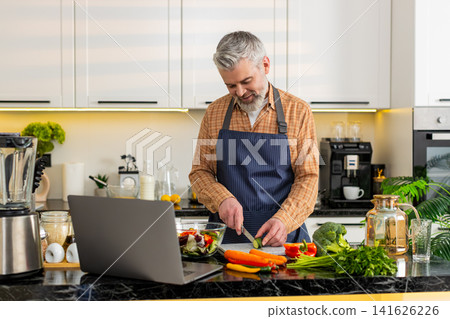 Middle-aged man preparing vegetable salad using laptop recipe in home kitchen for healthy diet meal Middle-aged man preparing vegetable salad using laptop recipe in home kitchen for healthy diet meal 141626226