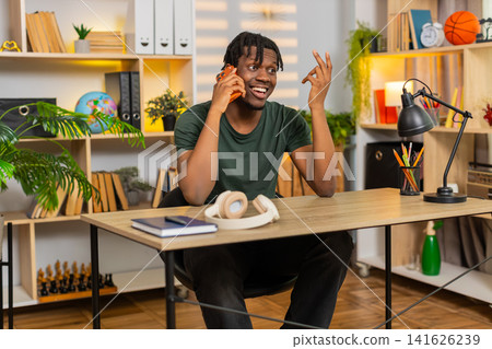 African American young man on home table answering smartphone call and smiling during chat indoors African American young man on home table answering smartphone call and smiling during chat indoors 141626239