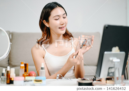 Asian beauty influencer filming a makeup tutorial using cosmetics, skincare products, smartphone and ring light in a modern home studio for social media content. Asian beauty influencer filming a makeup tutorial using cosmetics, skincare products, smartphone and ring light in a modern home studio for social media content. 141626432
