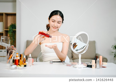 Asian woman practicing her daily skincare routine, gently touching her clear face while applying serum at a modern vanity. Asian woman practicing her daily skincare routine, gently touching her clear face while applying serum at a modern vanity. 141626448