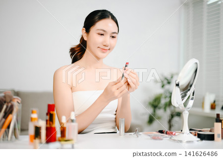 Young Asian woman applying makeup with eyeshadow palette while recording a beauty tutorial using smartphone and ring light in a modern home studio. Young Asian woman applying makeup with eyeshadow palette while recording a beauty tutorial using smartphone and ring light in a modern home studio. 141626486