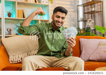 Indian man on home sofa counting cash and smiling after profit win or successful deal in living room Indian man on home sofa counting cash and smiling after profit win or successful deal in living room 141626831