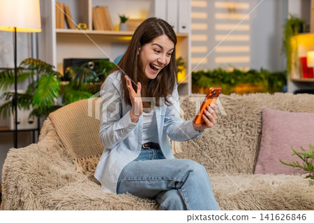 Young woman on home sofa with smartphone good news happy celebrating success in living room indoors Young woman on home sofa with smartphone good news happy celebrating success in living room indoors 141626846