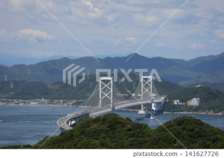 A landscape of cloudy skies, the Naruto Bridge, Awaji Island, the sea, ships sailing across the strait, the townscape along the coast of Shikoku, and a range of mountains. 141627726