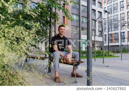 Man sitting on a bench outdoors and drinking coffee. Happy guy resting in city park near residential building. Casual urban lifestyle, break time in summer. Male enjoying drink. Man sitting on a bench outdoors and drinking coffee. Happy guy resting in city park near residential building. Casual urban lifestyle, break time in summer. Male enjoying drink. 141627882