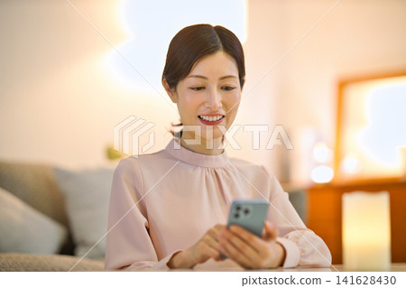 A woman in her 40s relaxing in her room at night and looking at her smartphone. A woman in her 40s relaxing in her room at night and looking at her smartphone. 141628430