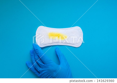 Blue Medical Gloved Hand Holding a Sanitary Pad with Yellow Urine Stains on Blue Background Blue Medical Gloved Hand Holding a Sanitary Pad with Yellow Urine Stains on Blue Background 141629360
