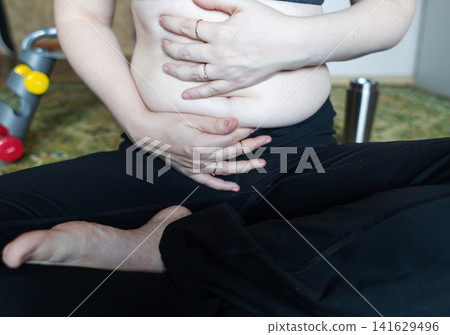 Woman demonstrating belly fat before doing yoga for weight loss and breathing exercises. Concept of body positivity, self love, and home fitness. Woman demonstrating belly fat before doing yoga for weight loss and breathing exercises. Concept of body positivity, self love, and home fitness. 141629496
