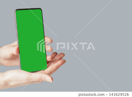 A hand holds a mockup phone with a bright green chroma key screen against a plain blue-grey A hand holds a mockup phone with a bright green chroma key screen against a plain blue-grey 141629526