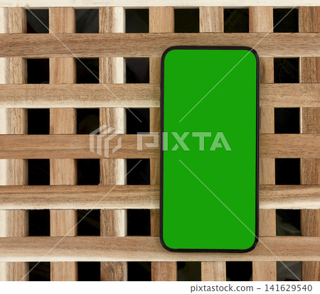 A phone with a green chroma key screen sits vertically on a dark wooden lattice pattern. The mobile A phone with a green chroma key screen sits vertically on a dark wooden lattice pattern. The mobile 141629540