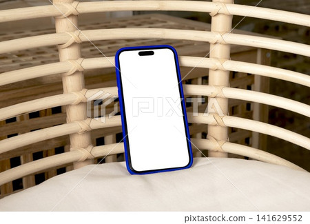 Blank smartphone mockup in blue case on a wicker chair. Angled mobile phone with copy space in an indoor lifestyle setting. App design and advertising template.. Blank smartphone mockup in blue case on a wicker chair. Angled mobile phone with copy space in an indoor lifestyle setting. App design and advertising template.. 141629552