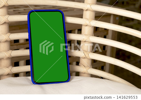 Blank smartphone mockup in blue case on a wicker chair. Angled mobile phone with green screen in an indoor interior. App design and advertising template with copy space.. Blank smartphone mockup in blue case on a wicker chair. Angled mobile phone with green screen in an indoor interior. App design and advertising template with copy space.. 141629553