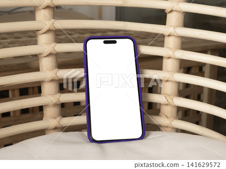 Blank smartphone mockup leaning on a woven rattan chair. Angled mobile phone with white screen in a cozy indoor setting. App design, advertising, and copy space template.. Blank smartphone mockup leaning on a woven rattan chair. Angled mobile phone with white screen in a cozy indoor setting. App design, advertising, and copy space template.. 141629572