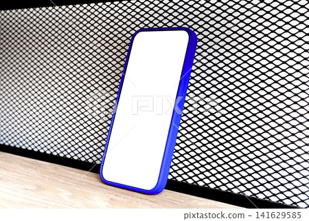 Blue-framed phone mockup with a blank white screen stands upright against a black wire mesh shelf. Blue-framed phone mockup with a blank white screen stands upright against a black wire mesh shelf. 141629585