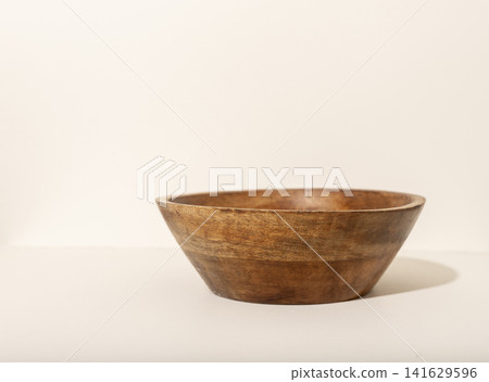 A rustic wooden bowl sits on a light beige surface photographed from a low side angle. The handcrafted bowl casts a soft shadow revealing its natural form and texture. Concept of 141629596