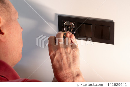 Older person's hand works on a black electrical socket frame on a white wall. The senior electrician Older person's hand works on a black electrical socket frame on a white wall. The senior electrician 141629641