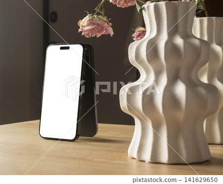 Phone mockup with a blank white screen stands on a wooden table beside decorative ceramic vases and Phone mockup with a blank white screen stands on a wooden table beside decorative ceramic vases and 141629650