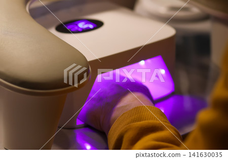 A person curing gel nail polish under a professional UV LED lamp in a beauty salon, illustrating manicure procedures, nail care, and cosmetic treatments A person curing gel nail polish under a professional UV LED lamp in a beauty salon, illustrating manicure procedures, nail care, and cosmetic treatments 141630035