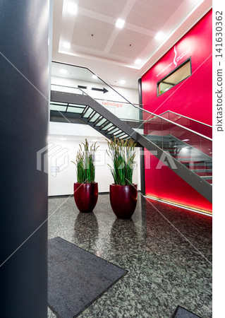 Modern office lobby with red feature wall and glass staircase, contemporary corporate interior with granite floor, cylindrical columns and large plant pots Modern office lobby with red feature wall and glass staircase, contemporary corporate interior with granite floor, cylindrical columns and large plant pots 141630362