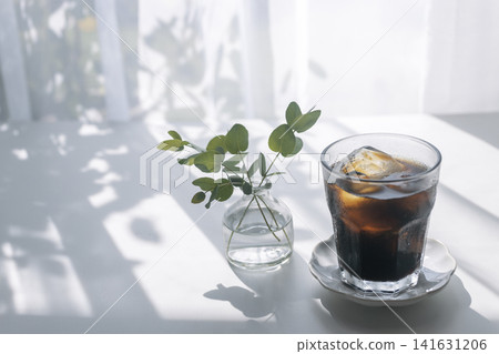 一張溫馨的咖啡館照片,桌上放著一杯冰咖啡,陽光斑駁地灑在桌上,周圍環繞著尤加利樹葉。 一張溫馨的咖啡館照片,桌上放著一杯冰咖啡,陽光斑駁地灑在桌上,周圍環繞著尤加利樹葉。 141631206