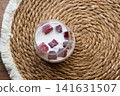 Homemade iced coffee prepared with frozen cherry juice ice cubes in a clear glass. Refreshing summer drink served on a natural woven placemat with soft daylight and minimal home interior background. 141631507