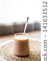 Homemade iced coffee prepared with frozen cherry juice ice cubes in a clear glass. Refreshing summer drink served on a natural woven placemat with soft daylight and minimal home interior background. 141631512