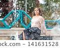 Young Woman Enjoys Relaxing Moment by Fountain in City Park on a Sunny Day 141631528