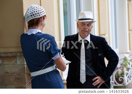 The conversation between the elderly stylish man and the woman in vintage clothes near the building on a sunny day The conversation between the elderly stylish man and the woman in vintage clothes near the building on a sunny day 141633026
