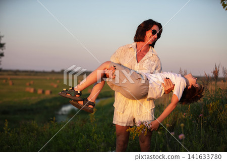 A joyful summer moment with a woman holding the child on the field during sunset. A joyful summer moment with a woman holding the child on the field during sunset. 141633780