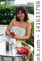 A woman is relaxing on an open terrace of a cafe, talking on the phone, next to a cup and saucer, a teapot and a flower arrangement in white, green, pink and silver tones, front view, lifestyle and 141633798